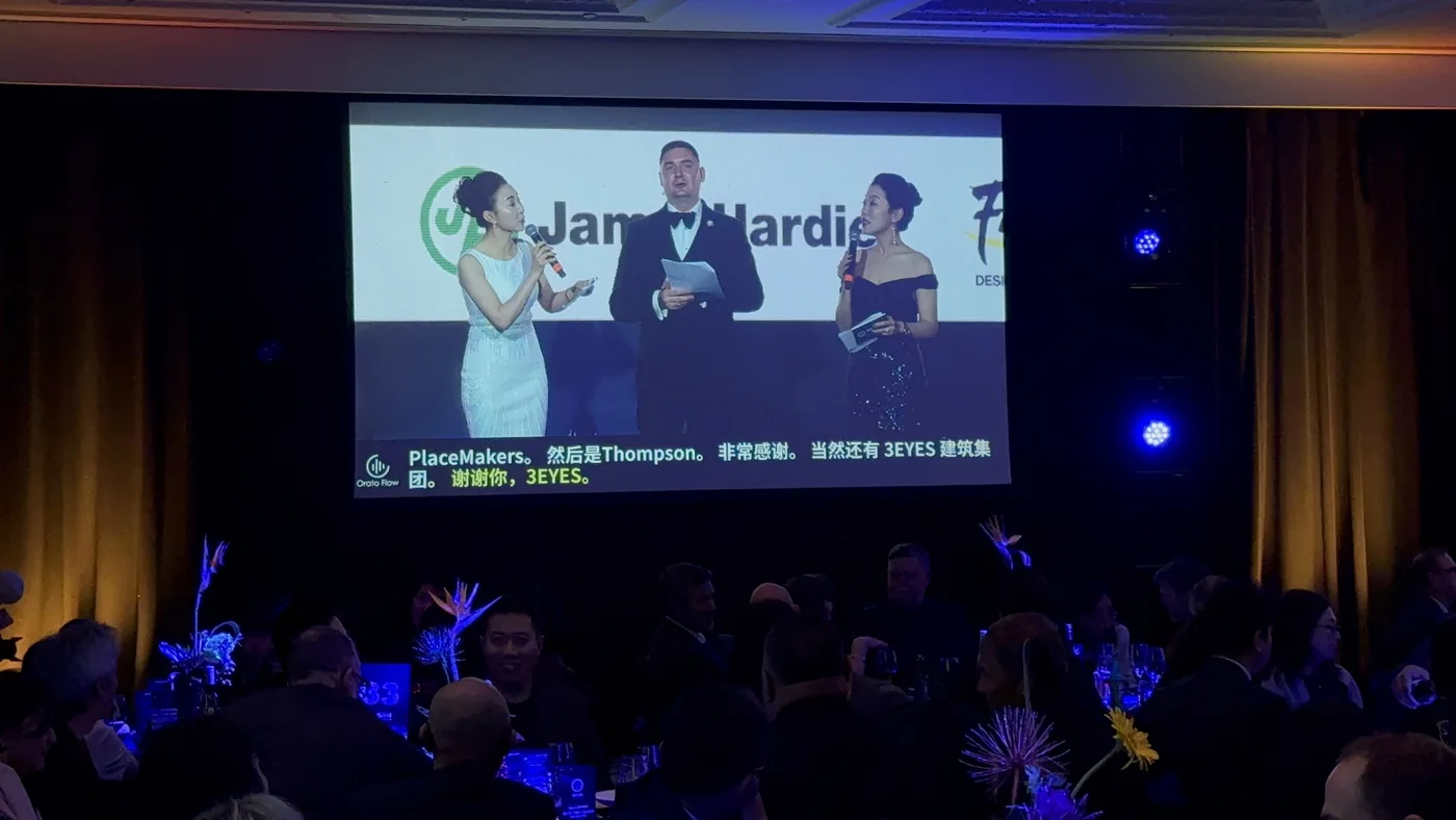 Three MCs on the main screen at the NZCBIA Excellence Awards with Orato Flow’s live Chinese captions beneath them, sponsor and company names left in English.