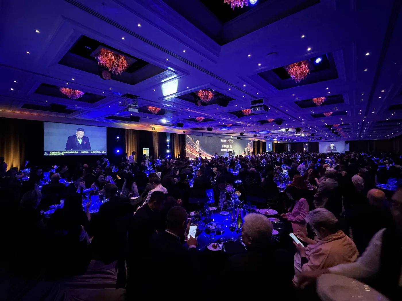 Guests at their tables watching the association president’s address on the main screen, with live Chinese captions along the bottom.