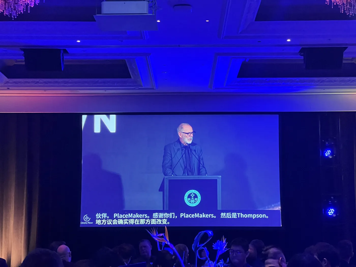 A speaker at the NZCBIA lectern on the main screen with live Chinese captions beneath, brand names such as PlaceMakers still rendered in English.