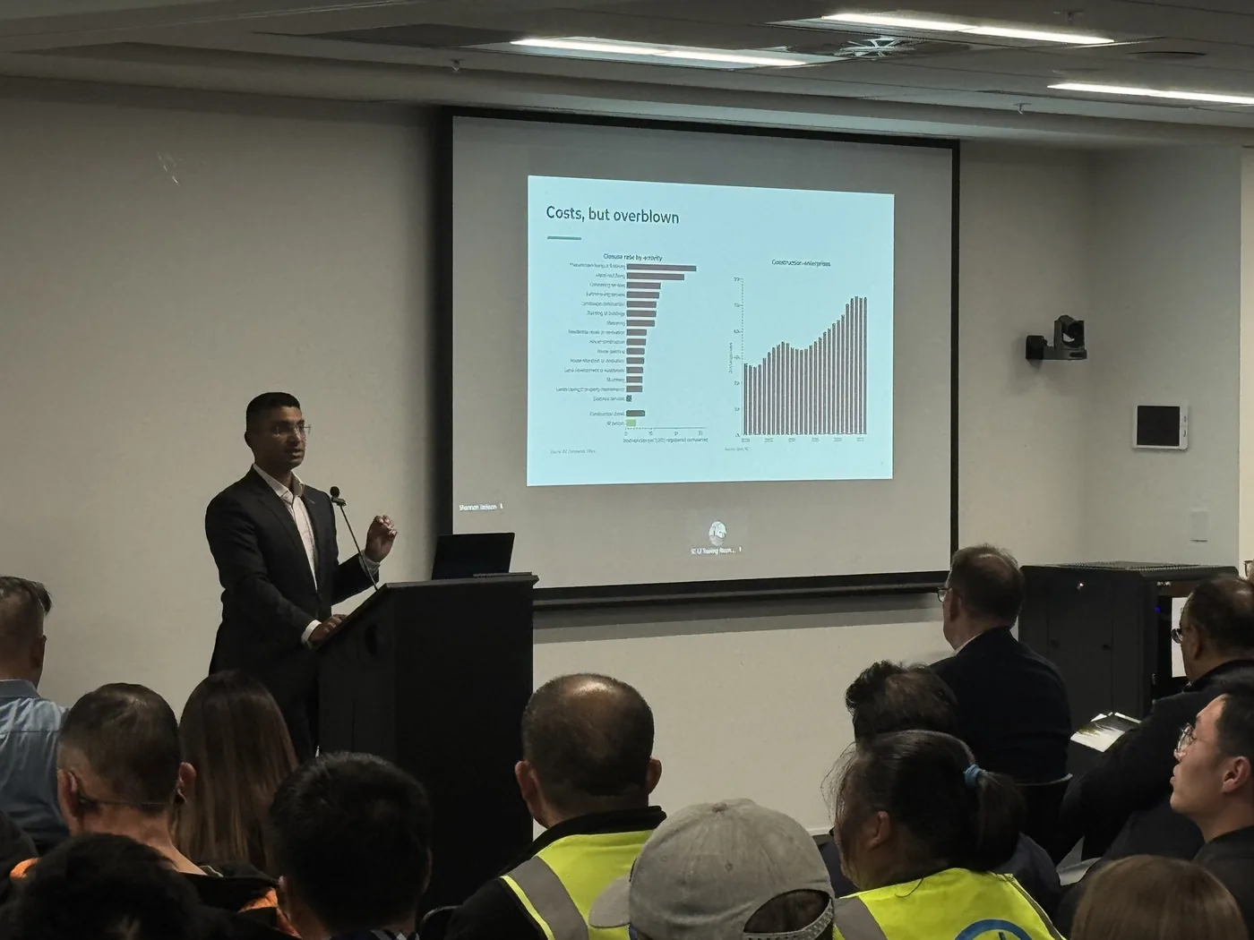 A presenter at the lectern with a chart slide from the 2026 NZCBIA Building & Construction Report on the projector screen, in front of a seated audience.