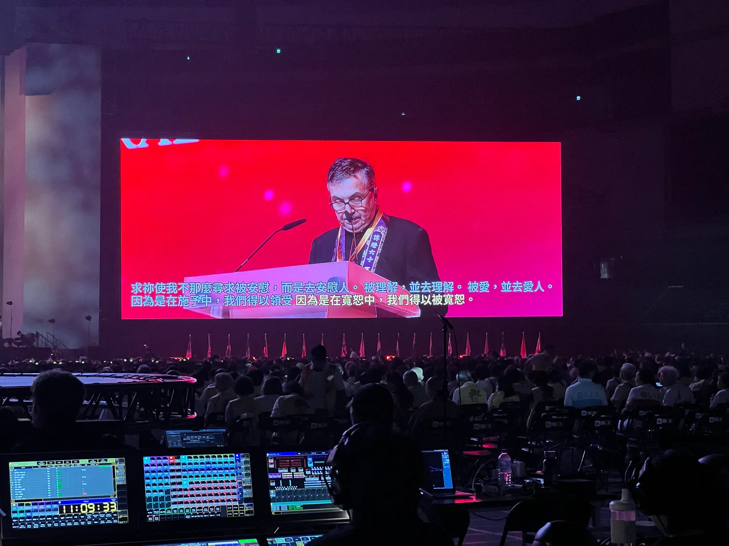 A guest speaker at a lectern on one of the Taipei Dome screens with live Chinese captions beneath, seen from behind the production desk’s consoles.