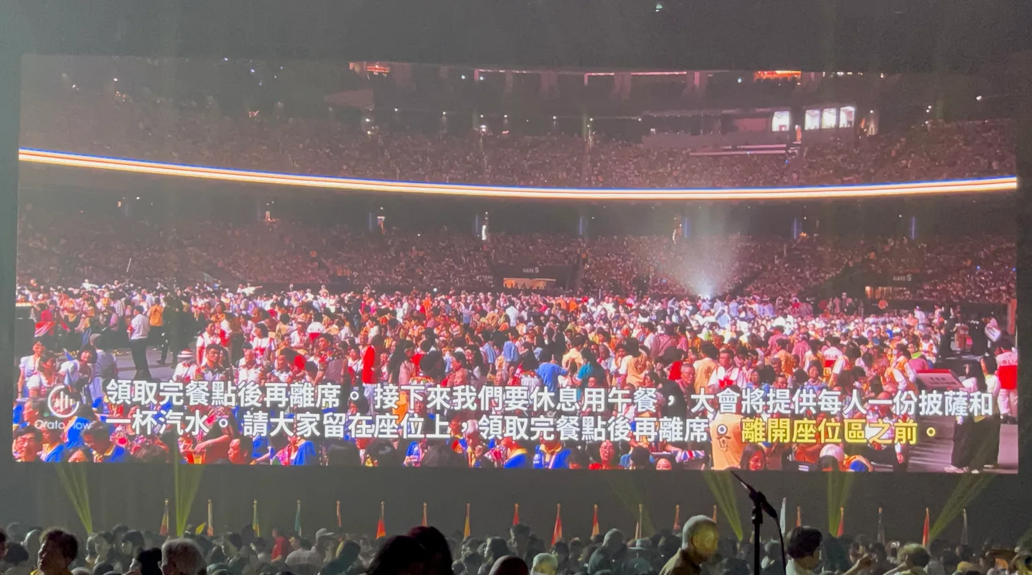 Two lines of live Chinese captions along the bottom of a Taipei Dome screen explaining the lunch arrangements, over a camera shot of the crowded arena floor.