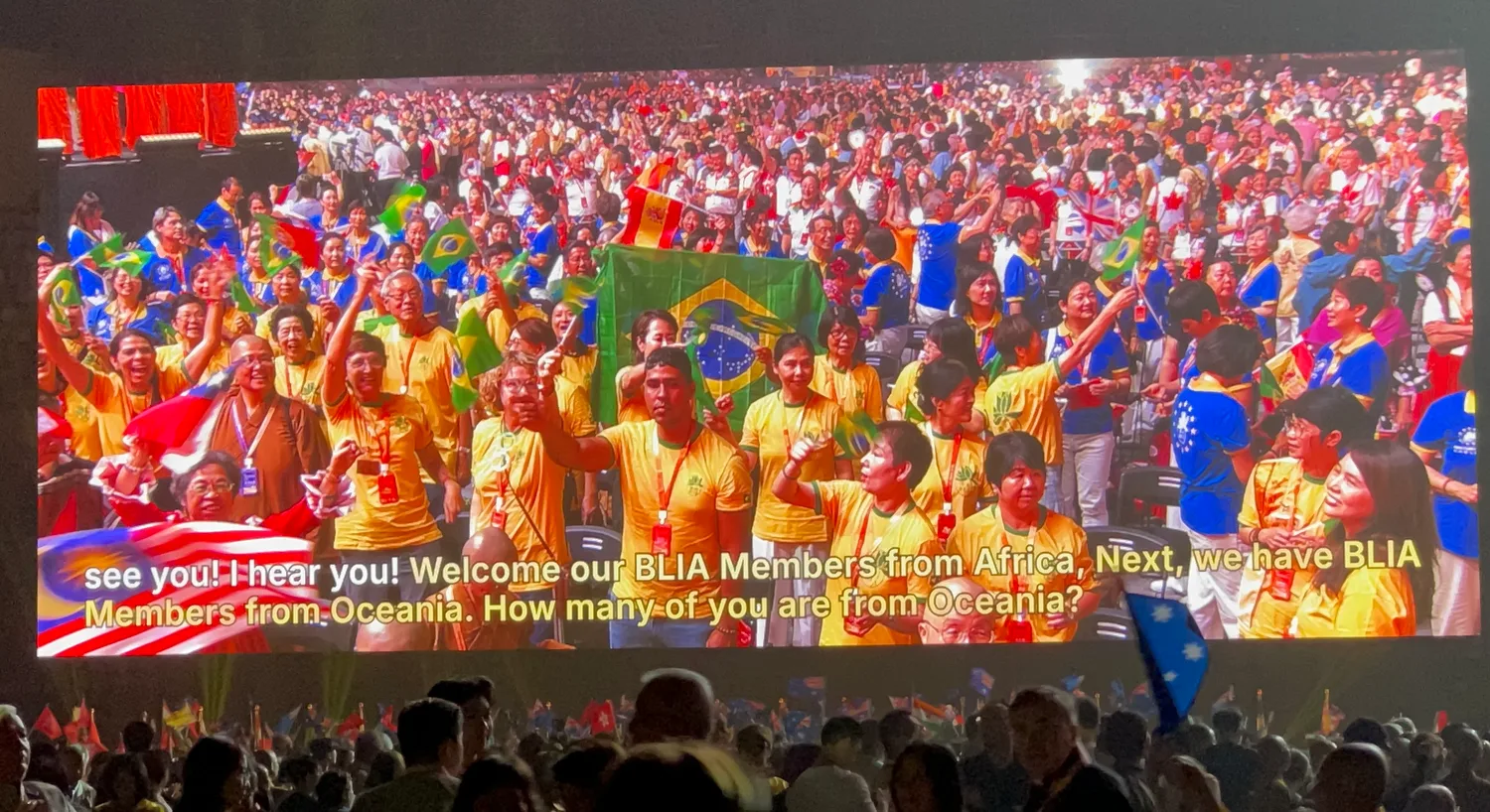 English captions along the bottom of a Taipei Dome screen as the host welcomes members region by region, over a live camera shot of the crowd waving national flags.